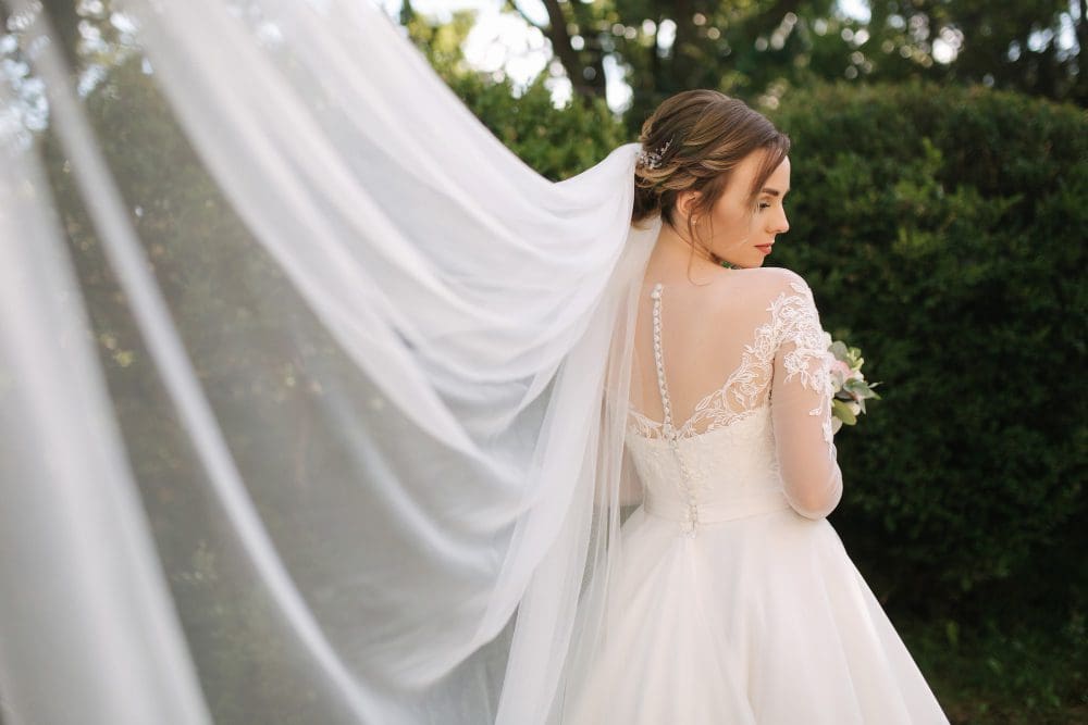 A bride in a long-sleeved lace wedding dress stands outdoors, looking over her shoulder, with her veil flowing behind her.