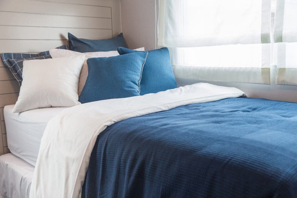 A neatly made bed with a blue blanket, white sheets, and several pillows, positioned next to a window with sheer curtains letting in natural light.