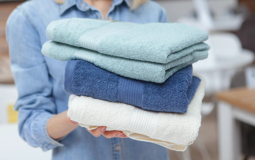A person in a denim shirt holds three folded towels in light blue, navy blue, and white.