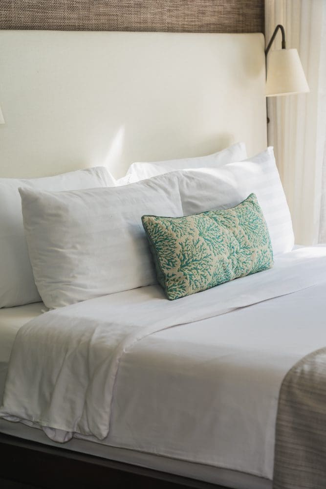 A neatly made bed with white linens, two white pillows, and a decorative pillow with a green coral pattern, positioned next to a wall lamp.