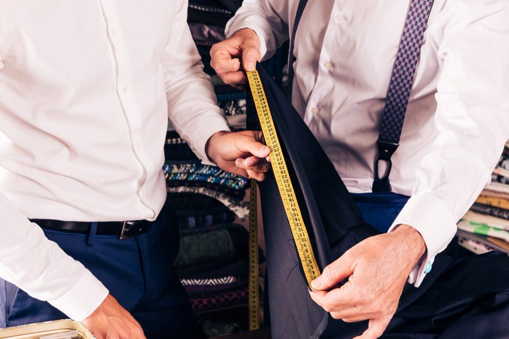 Two men in dress shirts measure a pair of pants with a yellow measuring tape in a fabric store or tailor shop.