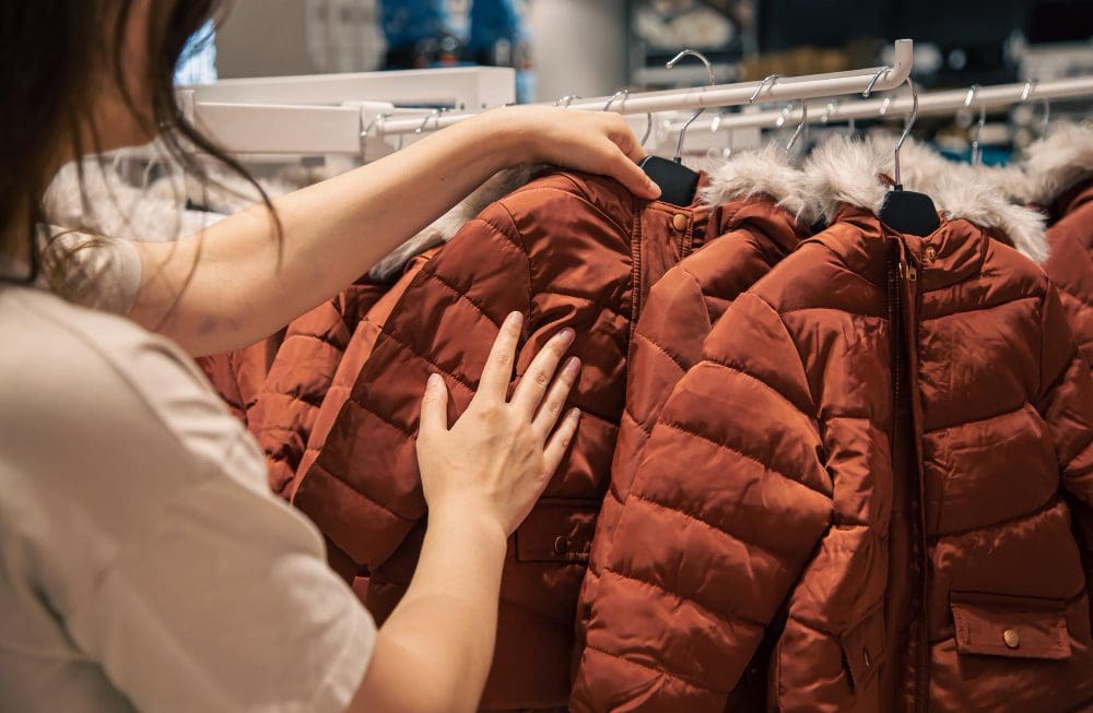 A person examines red winter jackets with fur-trimmed hoods hanging on a clothing rack in a store.