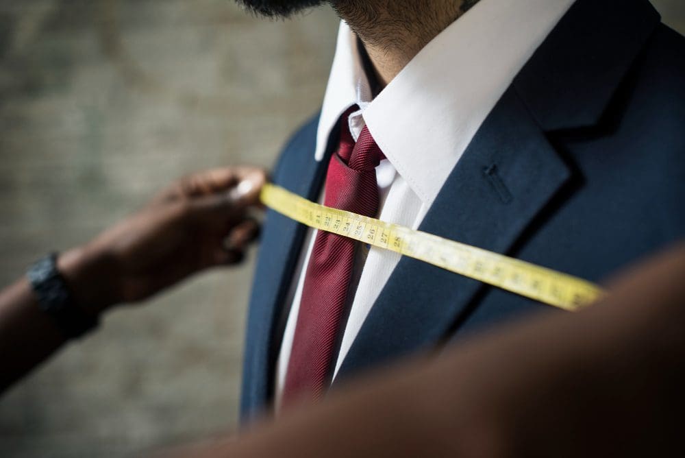 A person in a suit and tie is having their chest measured with a yellow measuring tape by another person.