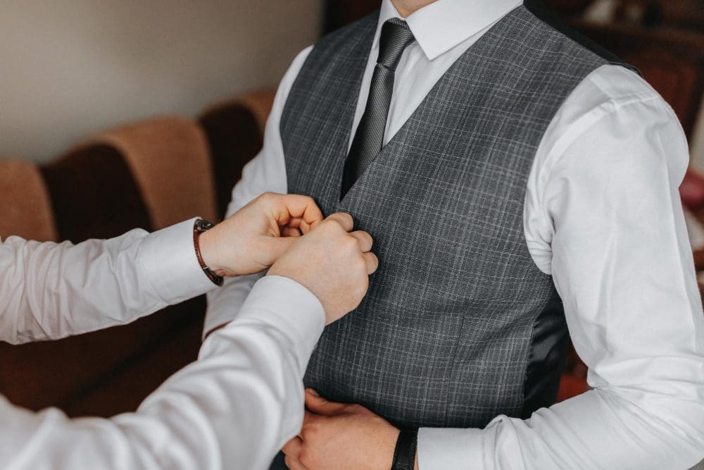 A person is buttoning the vest of a man dressed in a white shirt, grey checkered vest, and black tie, preparing him for a formal event.