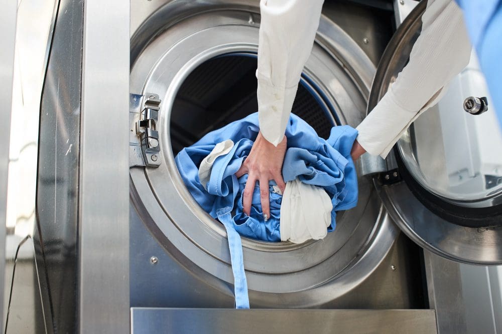 A person loads blue and white laundry into an industrial washing machine.