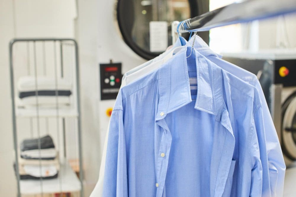 A close-up of freshly cleaned blue shirts on hangers at a dry cleaning facility, with laundry machines and folded linens visible in the background.