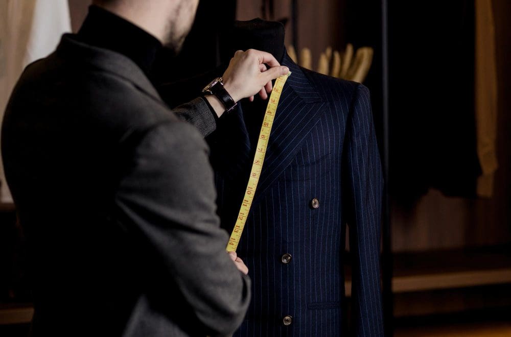 A tailor measures the front of a blue pinstripe suit jacket on a mannequin using a yellow measuring tape.