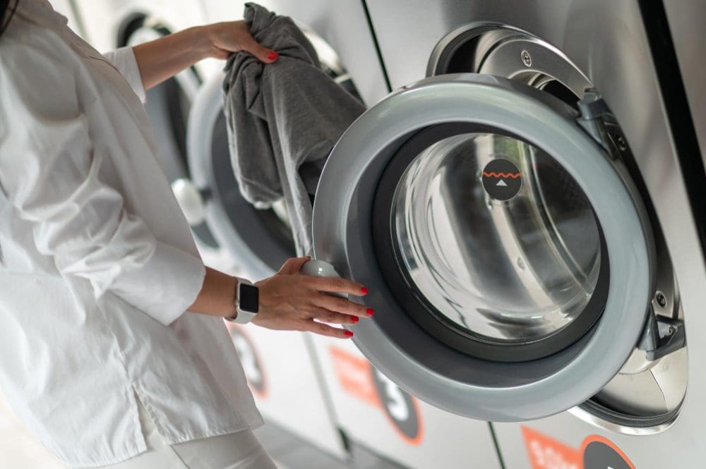 Person placing laundry into a front-loading washing machine at a laundromat, wearing a white shirt and a smartwatch.