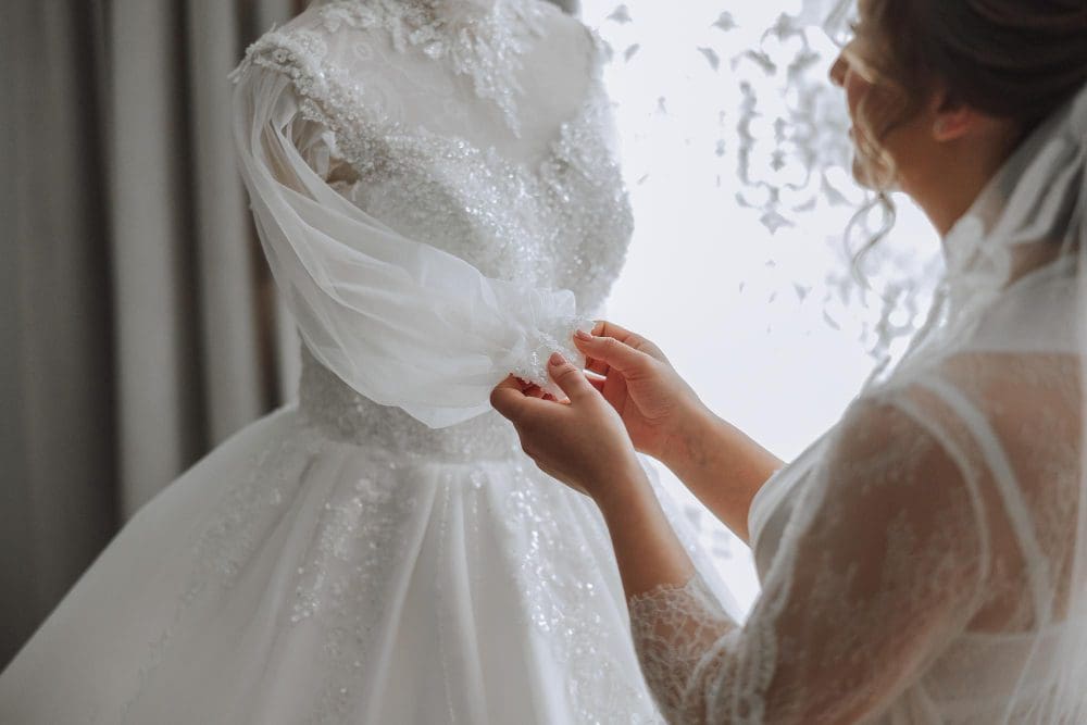 A bride wearing a lace robe adjusts the sleeve of a white, sequined wedding dress on a mannequin near a window.