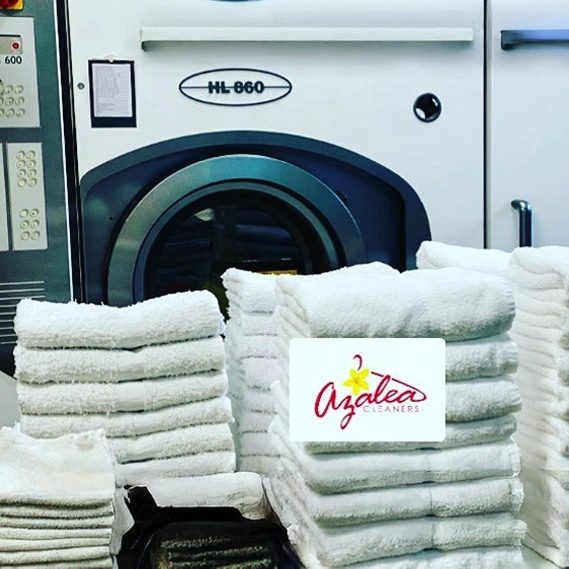 Stacks of folded white towels are arranged in front of a large industrial washing machine, with an Azalea Cleaners sign placed on top of one pile.