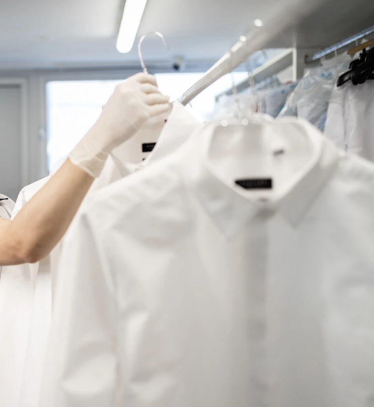 A person wearing a white glove is hanging or inspecting white dress shirts on hangers in a well-lit room.
