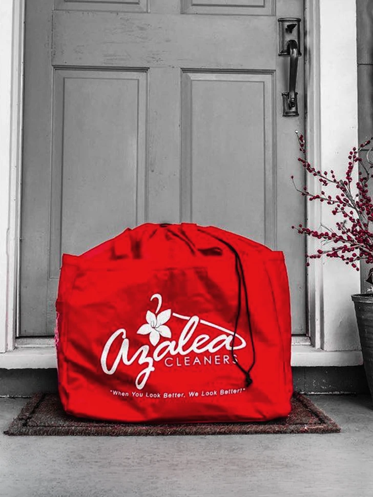 A large red laundry bag labeled "Azalea Cleaners" sits on a doormat in front of a closed door with a vase of red branches nearby.
