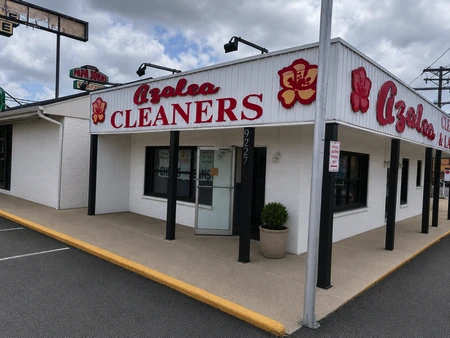 A white building with red "Azalea Cleaners" signage, a hibiscus flower logo, glass front door, and paved parking area under a cloudy sky.